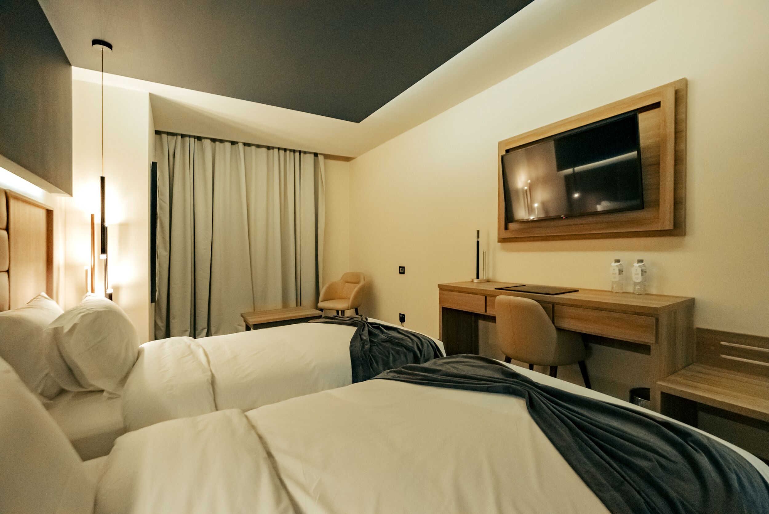 A hotel room with two beds, a desk, a chair, a TV, and a large window with curtains.
