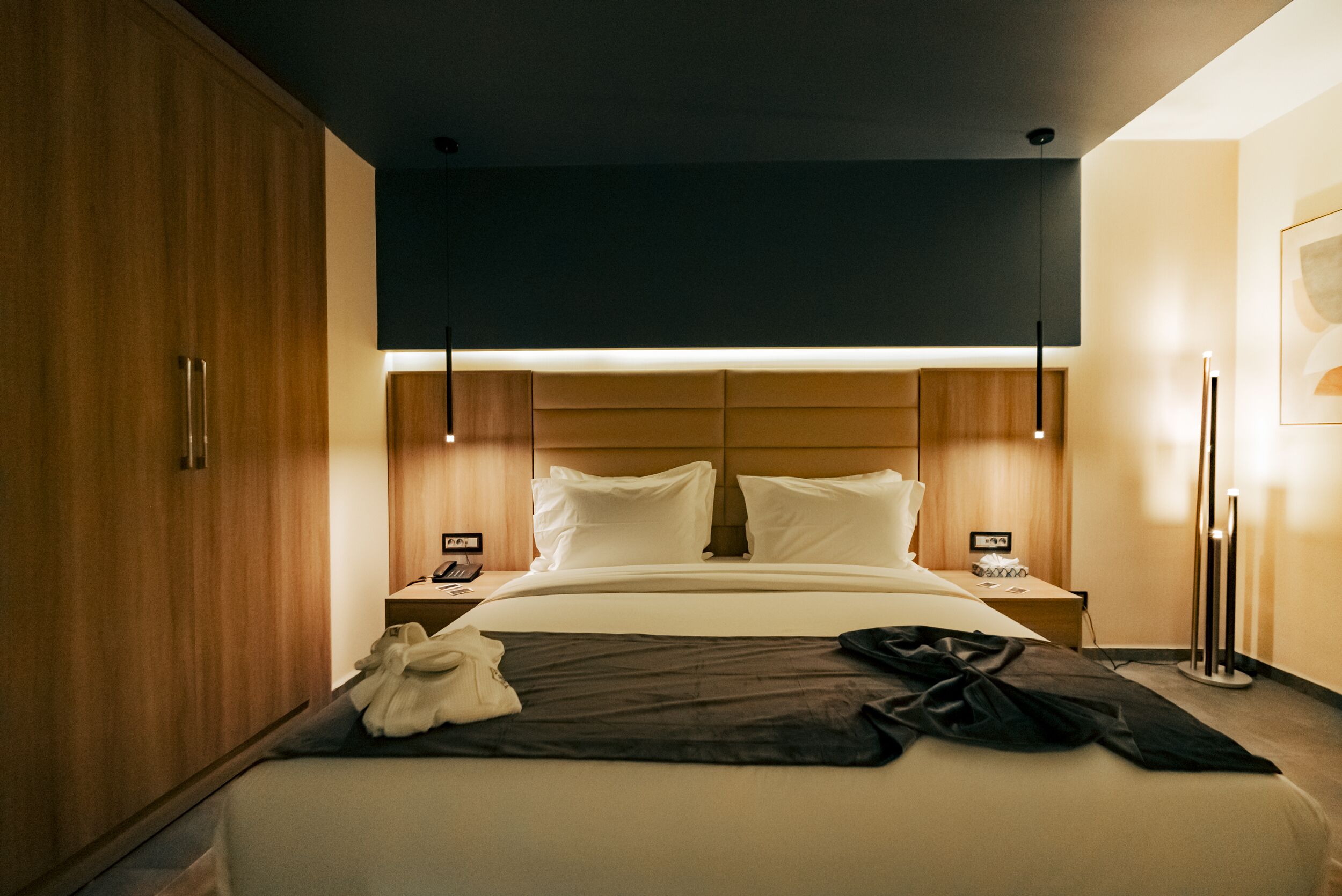 A hotel room with a large bed, wooden wardrobe, bedside tables, and modern pendant lighting.