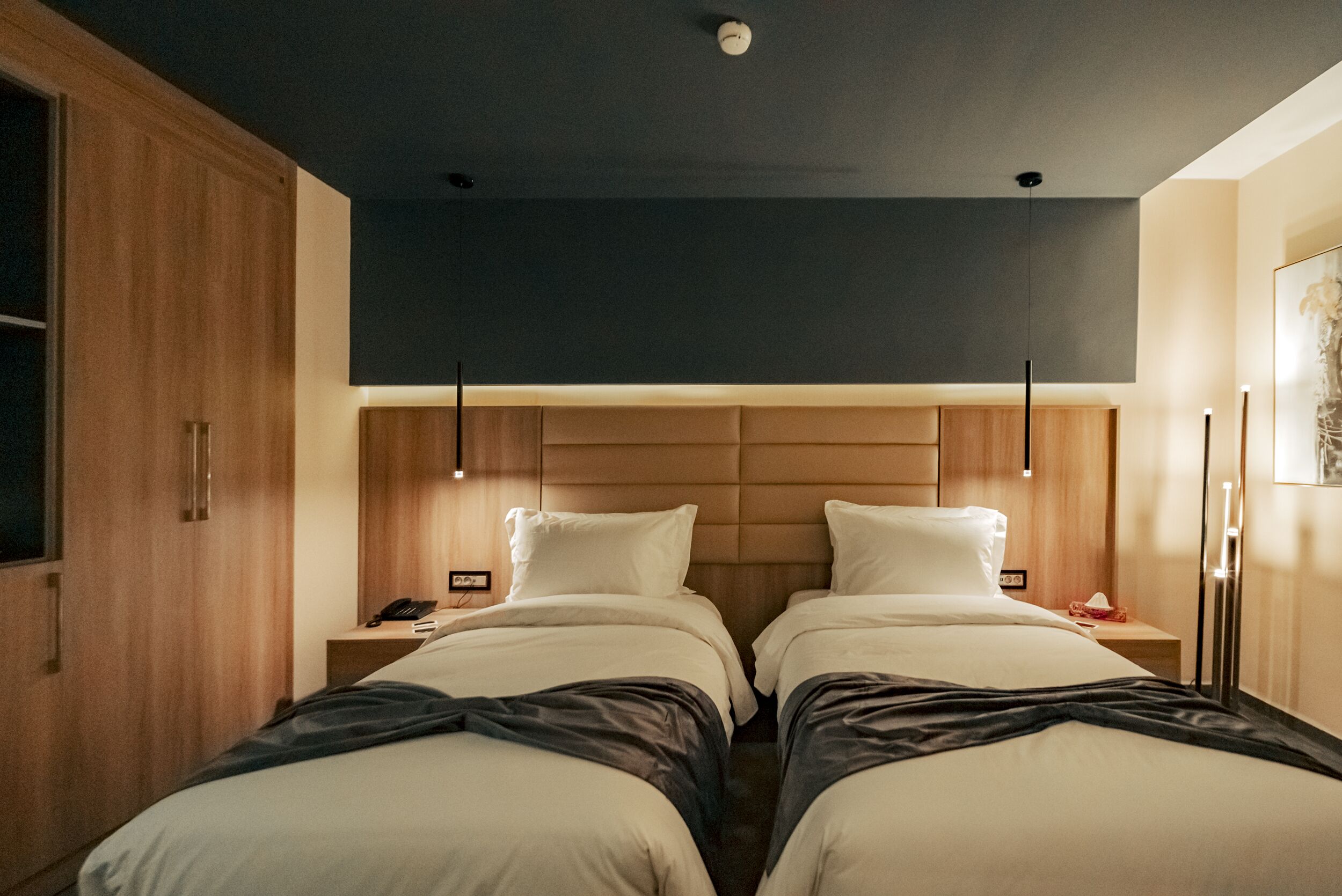 Ein Hotelzimmer mit zwei Betten, einem Holzschrank, Nachttischen mit Lampen und einem modernen Deckenstil.