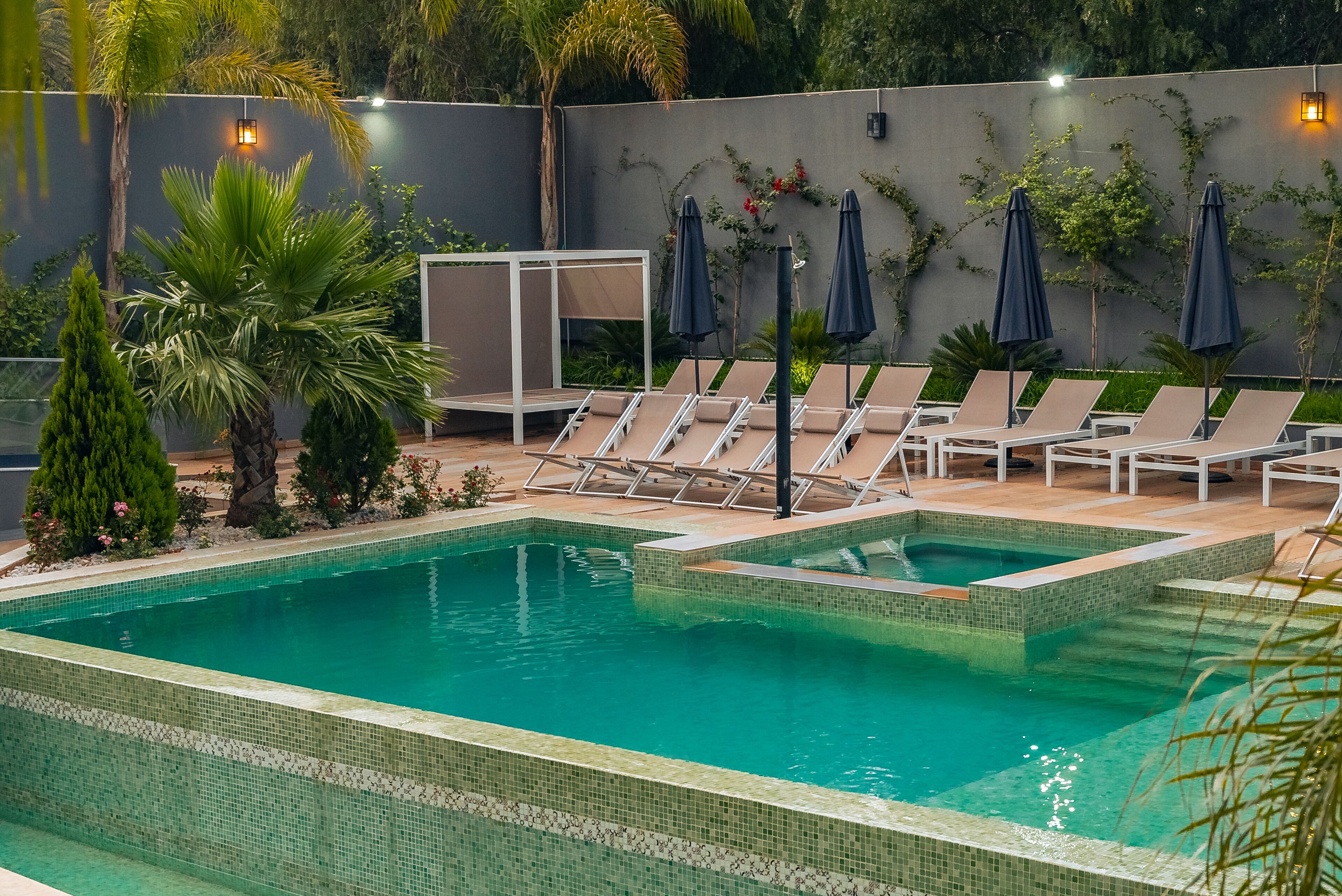 Ein rechteckiger Pool mit gefliesten Rändern, umgeben von Liegestühlen und Sonnenschirmen, mit einer Grünwand und einer Lampe.