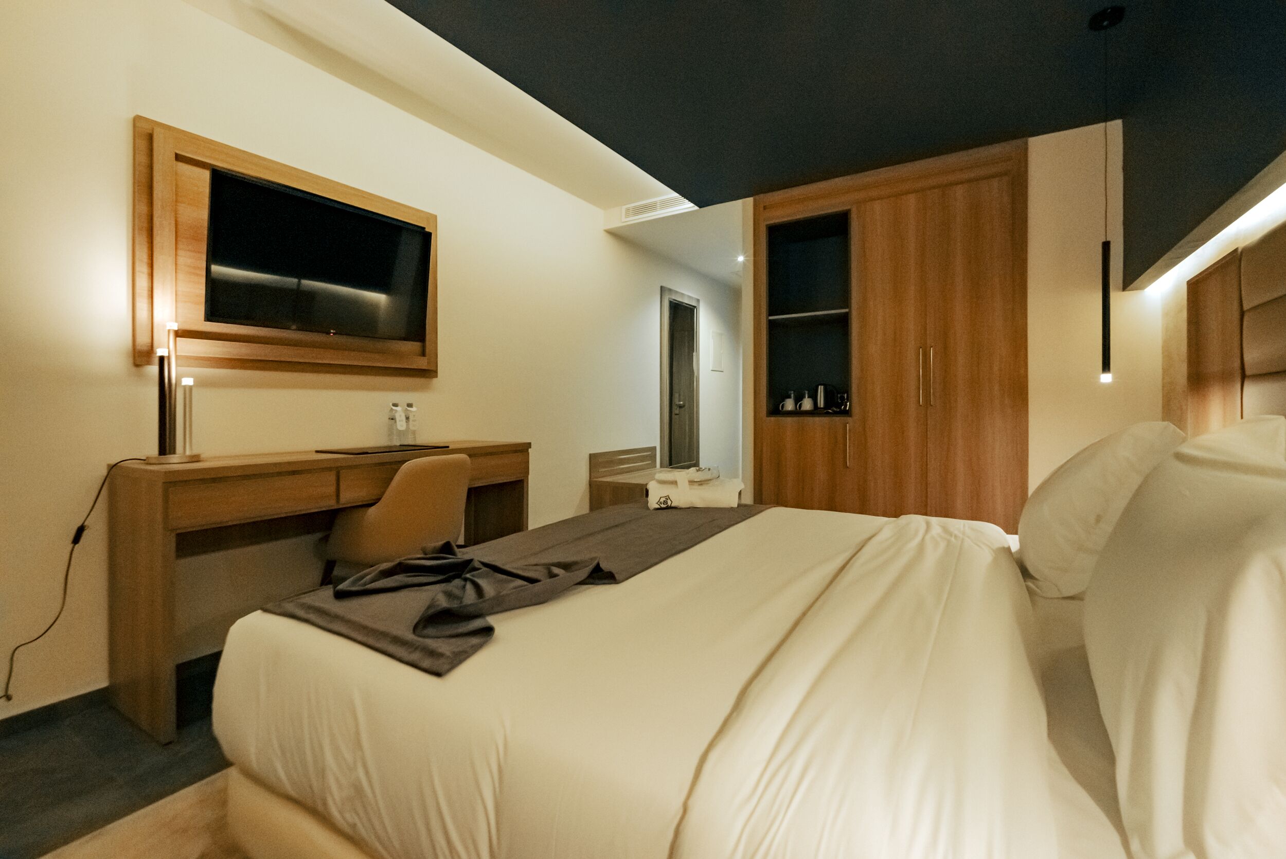 A modern hotel room with a bed, desk, chair, and built-in TV.