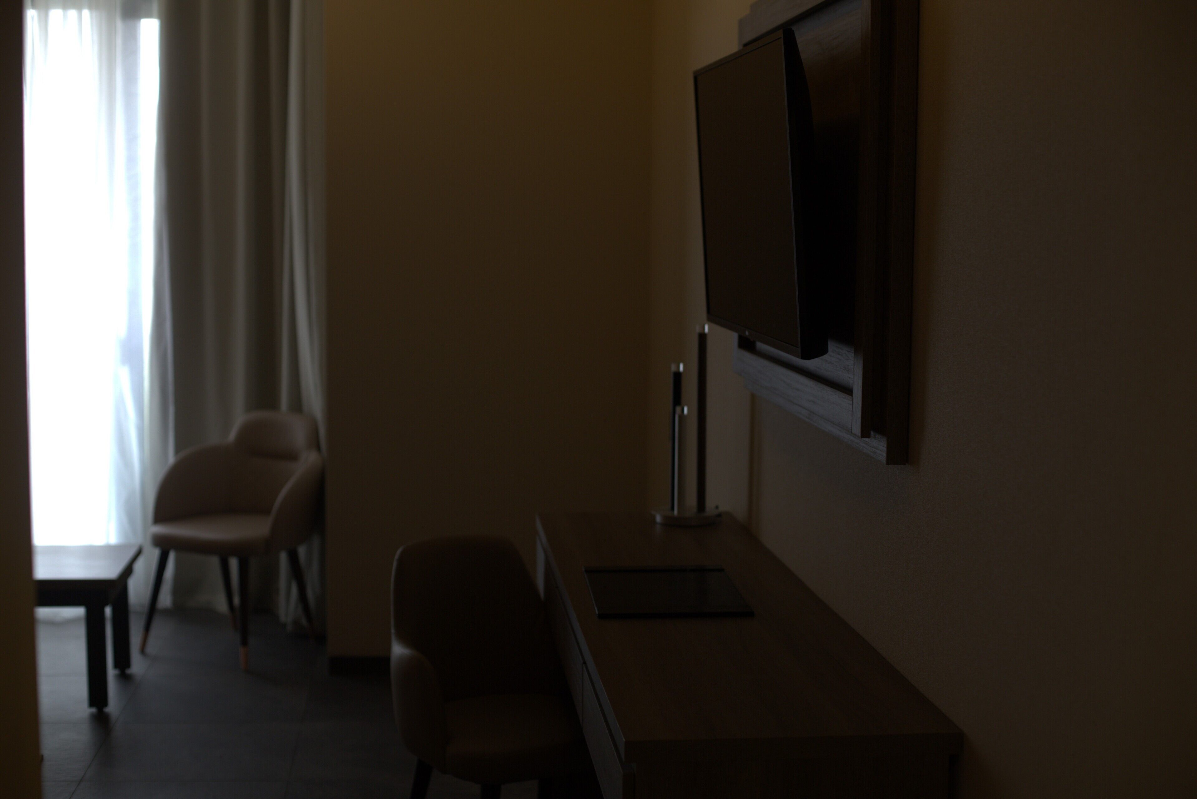 Una habitación de hotel tenuemente iluminada con escritorio, dos sillas, televisor y ventana con cortinas.