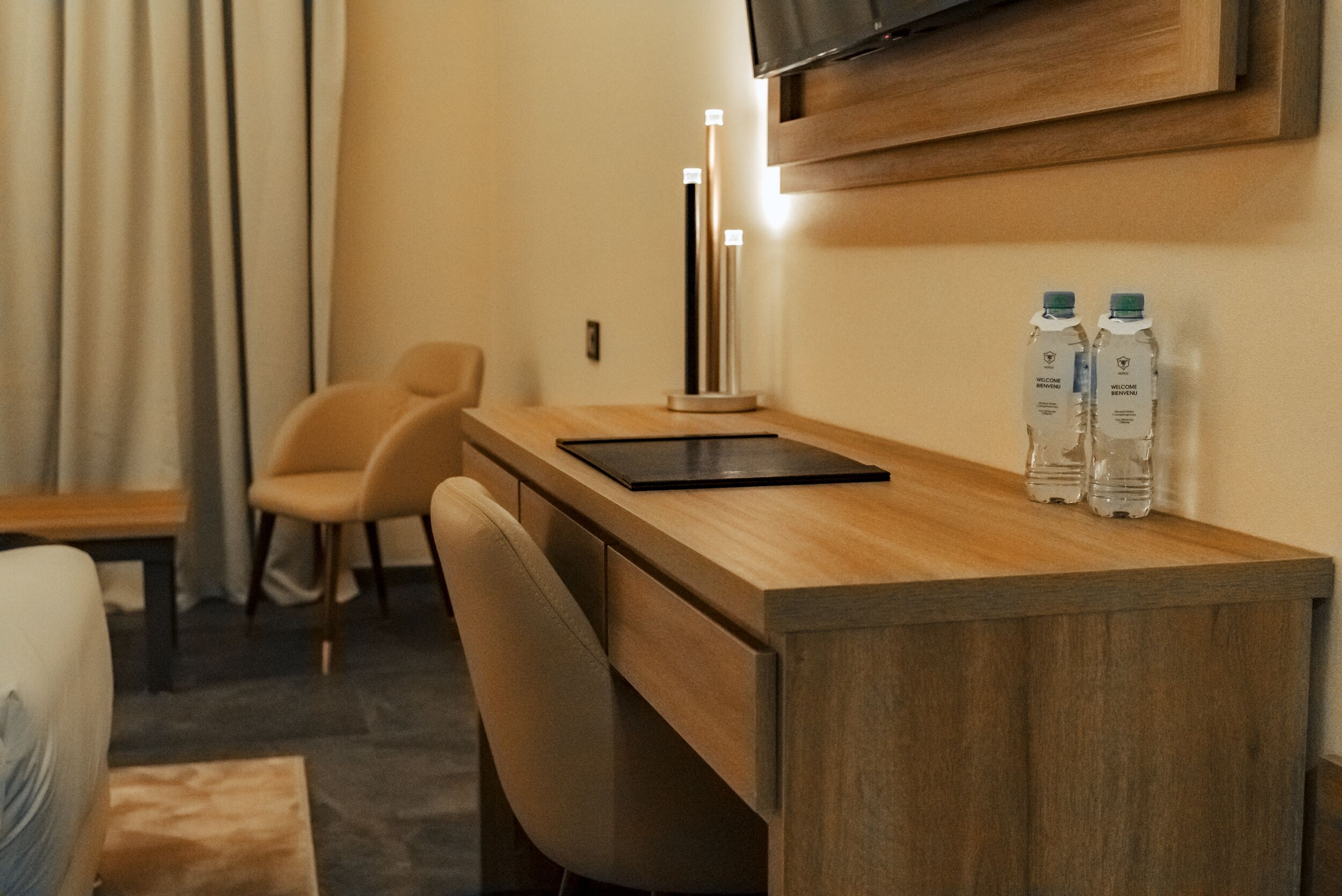 A hotel room with a wooden desk, a chair, a flat-screen TV mounted on the wall, and two bottles of water on the desk.