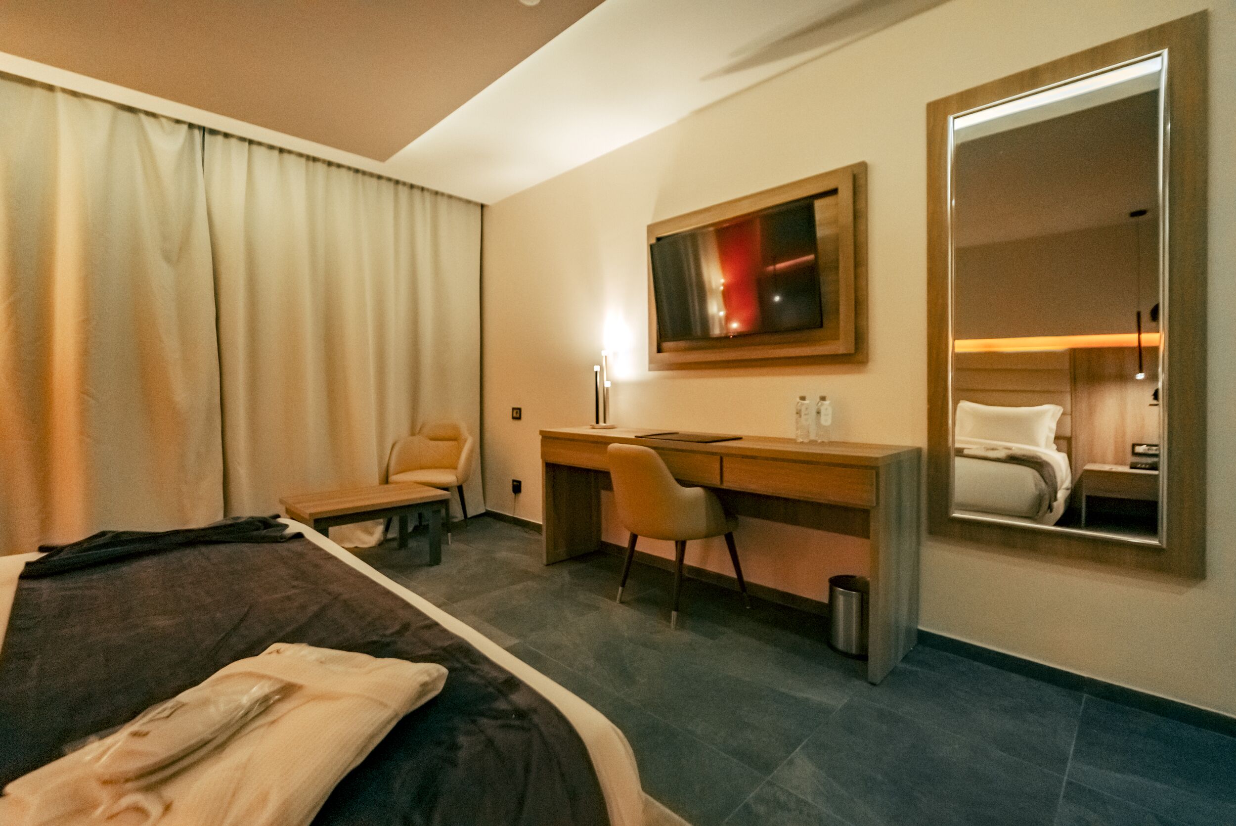 A hotel room with a bed, a desk, a chair, a mirror, and a window with curtains.