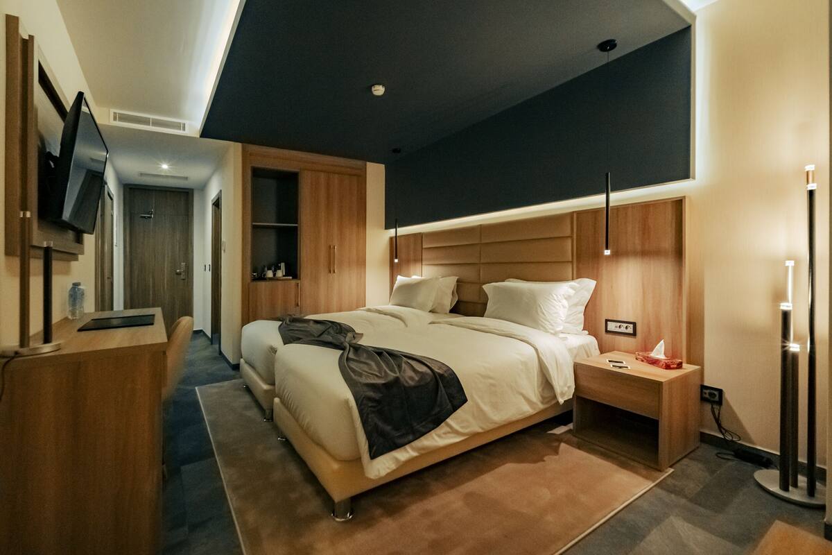 A modern hotel room with a large bed, bedside tables, a desk, a flat-screen TV, and a ceiling light fixture.