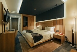 A modern hotel room with a large bed, bedside tables, a desk, a flat-screen TV, and a ceiling light fixture.