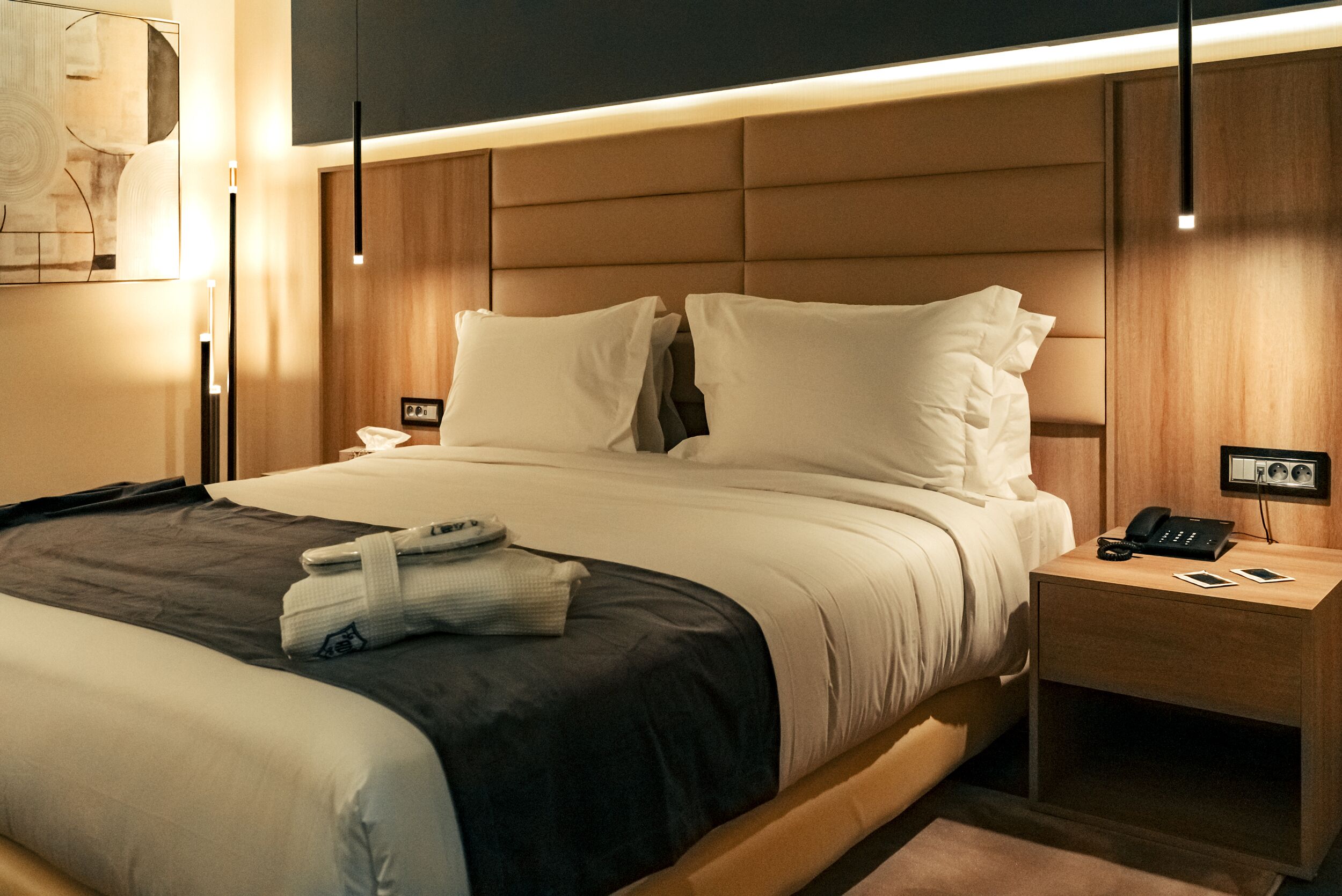 A modern hotel room with a large bed, a bedside table with a phone and a lamp, and a wall-mounted artwork.