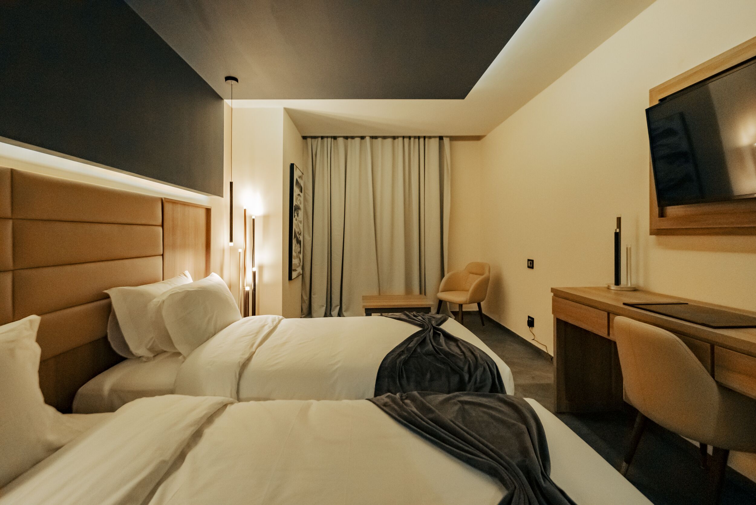 A hotel room with two beds, a desk, a chair, a TV, and a window with curtains.