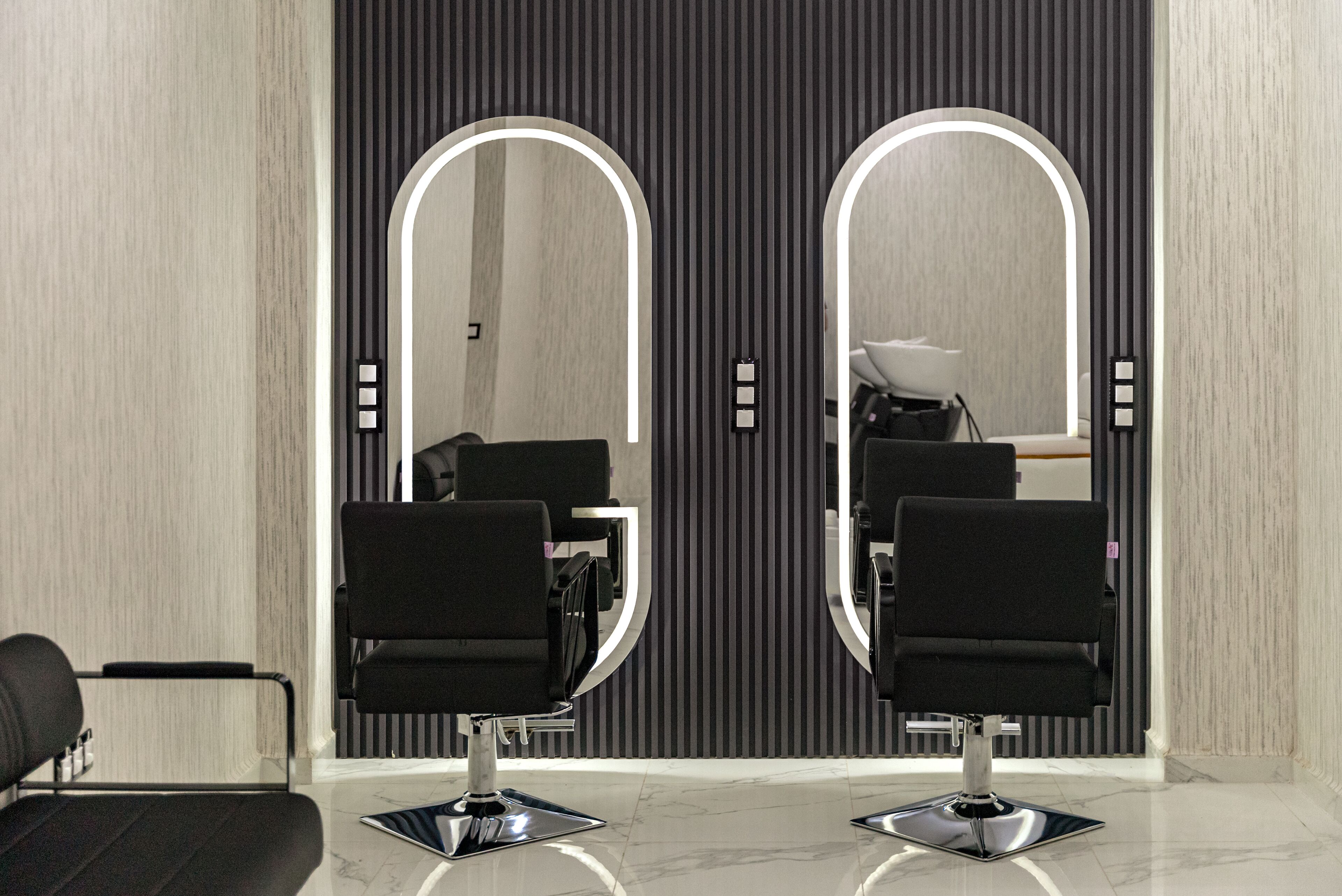 A modern salon with two black salon chairs, a large mirror with a lighted arch, and a hairdryer on a counter.