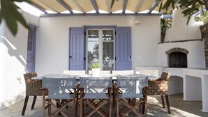 Un patio al aire libre con una mesa y sillas, piso de piedra y un edificio blanco con contraventanas azules.