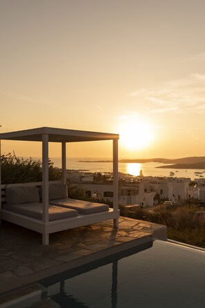 A sunlit outdoor seating area with a pool, overlooking a coastal cityscape during sunset. - Orasea Villa Paros (Paros)