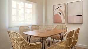 A dining area with a wooden table surrounded by wicker chairs, two abstract paintings on the wall, and a large window providing natural light. - Amador de los Rios Free Parking Gratis (Sevilla)