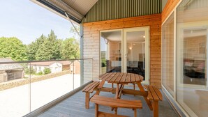 Eine Holzterrasse mit Tisch und Bänken, eine Glastür zu einem Innenraum und Blick auf eine ländliche Landschaft mit Bäumen und Gebäuden.
