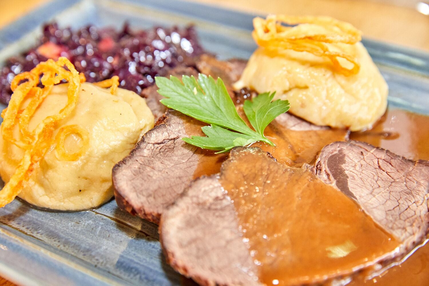 A plate of sliced beef with gravy, mashed potatoes, and a side of red cabbage garnished with fried onions and parsley.