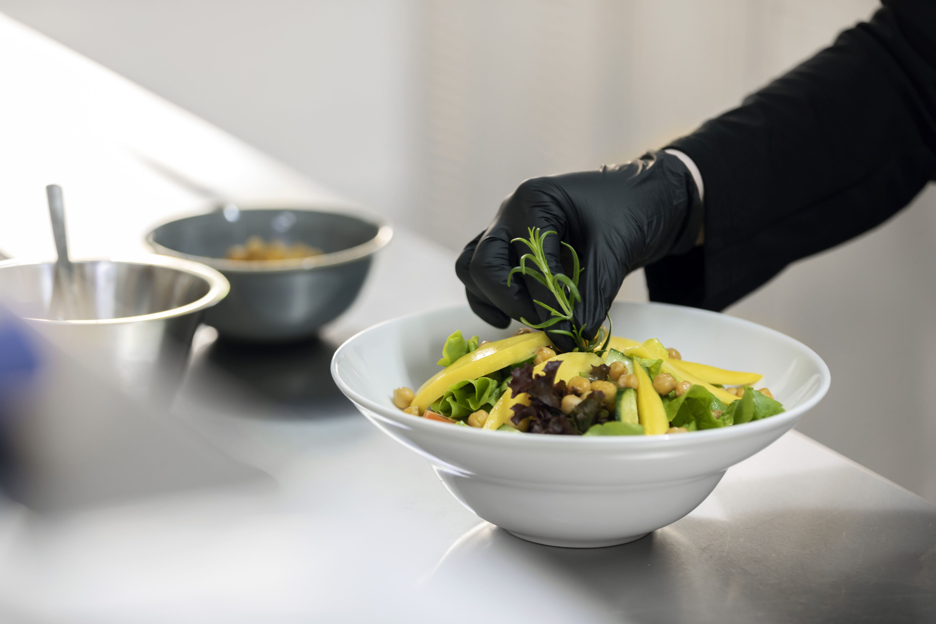 A person in a black glove adding a green herb to a white bowl of salad.