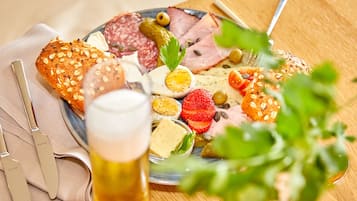 Une planche de charcuterie garnie de différentes sortes de viandes, de fromages et de fruits, accompagnée d’un verre de bière et de couverts sur une table en bois.