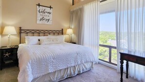 A bedroom with a bed, two bedside tables, a nightstand, a window with curtains, and a view of greenery outside. - Beautiful 6th floor, cathedral ceiling, ocean view condo (Pawleys Island)