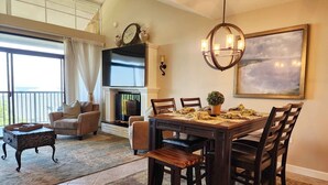 A dining area with a wooden table and chairs, a fireplace, and a large window with curtains. - Beautiful 6th floor, cathedral ceiling, ocean view condo (Pawleys Island)