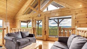 A wooden cabin interior with a large wooden ceiling, a sofa, a coffee table, and a view of green hills through large windows. - Aurora, Free Tickets, Breathtaking Views, “Cozy Cabin Retreat in the Smokies!!! (Sevierville)