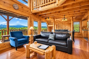 A wooden cabin interior with a sofa, armchair, and dining table, featuring large windows and a view of mountains.