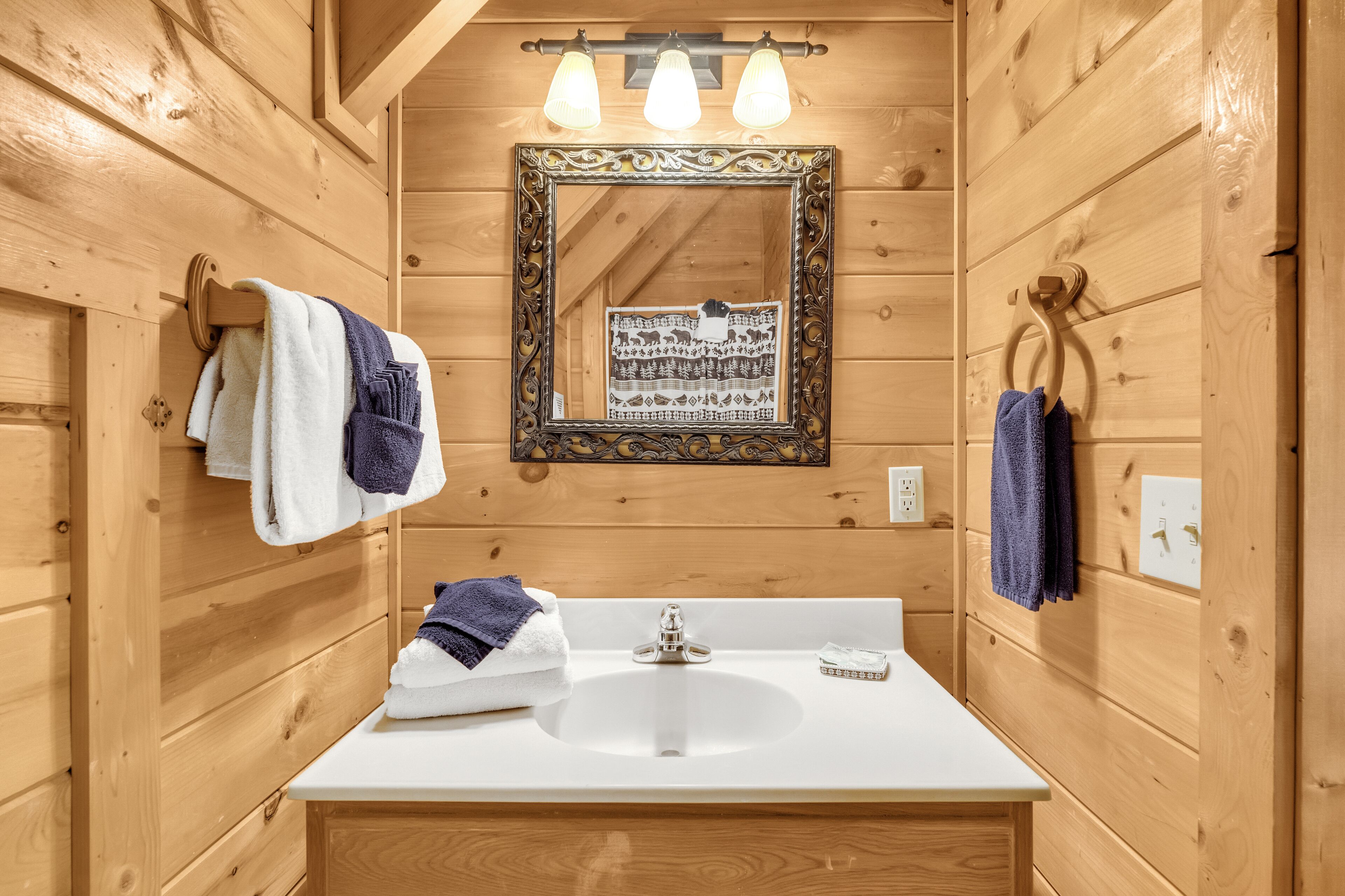 Un bagno in una cabina di legno con un lavandino bianco, tre asciugamani appesi e uno specchio con una cornice decorata.