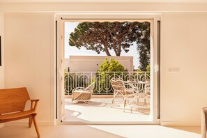 A room with a wooden bench, a white door leading to an outdoor balcony with a white railing, wicker chairs, and a table. - Maison Carla Rosa (Cannes)