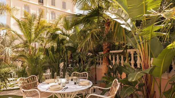 An outdoor dining area with wicker chairs and a round table set for four, surrounded by lush tropical plants. - Maison Carla Rosa (Cannes)