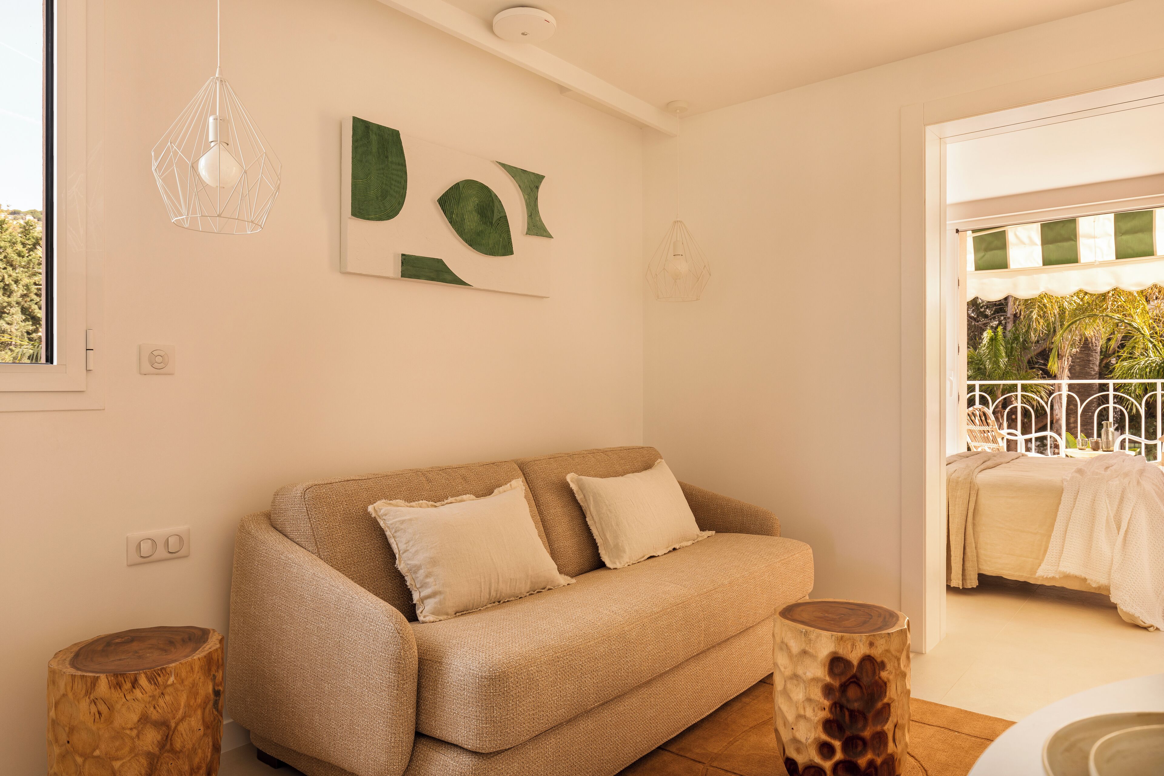 A cozy room with a beige sofa, two wooden side tables, and a view of a balcony with a bed.
