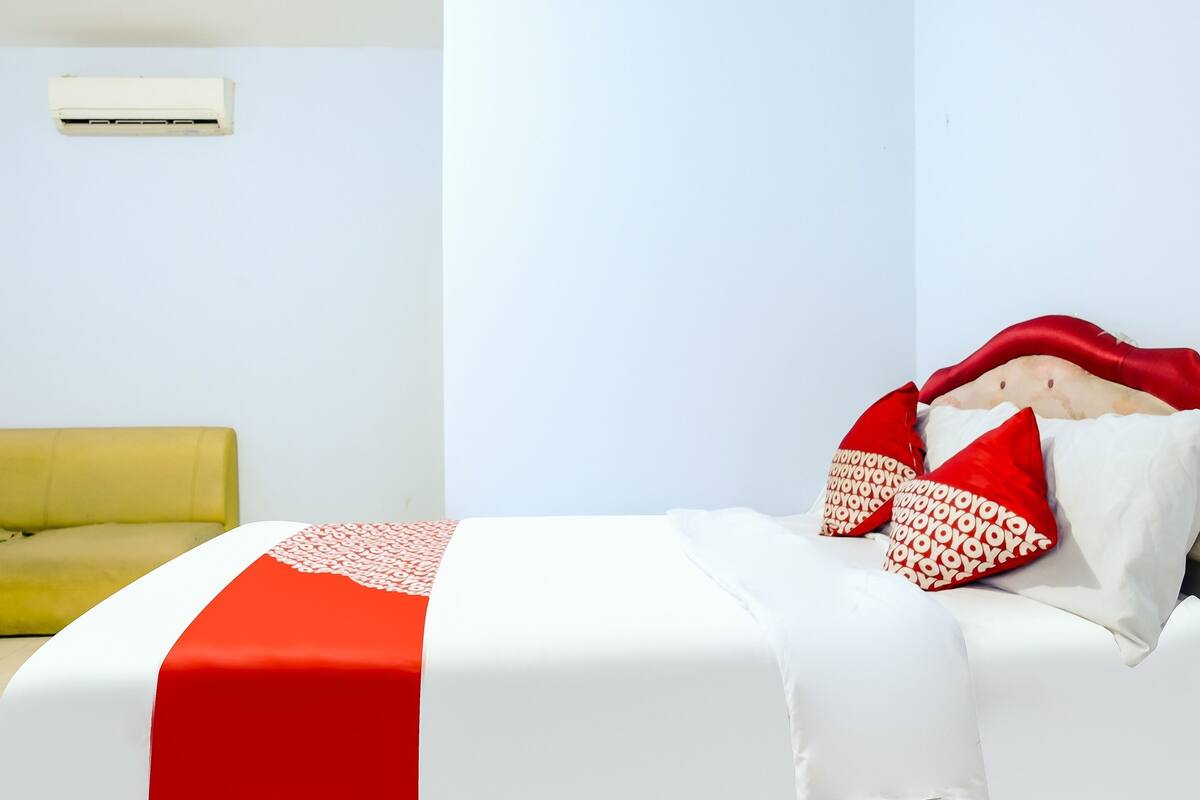 Un lit recouvert d’une couvre-lit rouge et blanche, un canapé jaune et un mur blanc sur lequel est installé un appareil de climatisation.