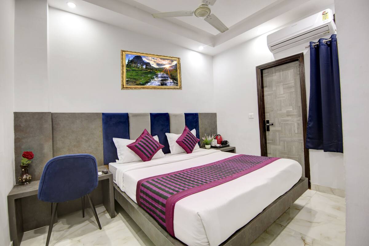 A bedroom with a bed, a desk, a chair, a nightstand, a wall-mounted air conditioner, and a door.