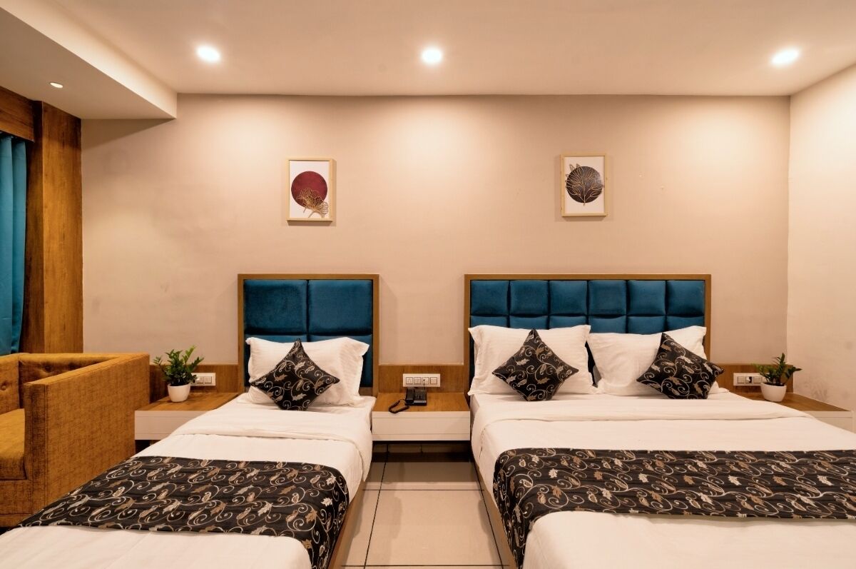 A hotel room with two beds, each with a blue headboard, white bedding, and patterned throw blankets. There are two bedside tables with lamps, a small potted plant, and a wall-mounted phone.