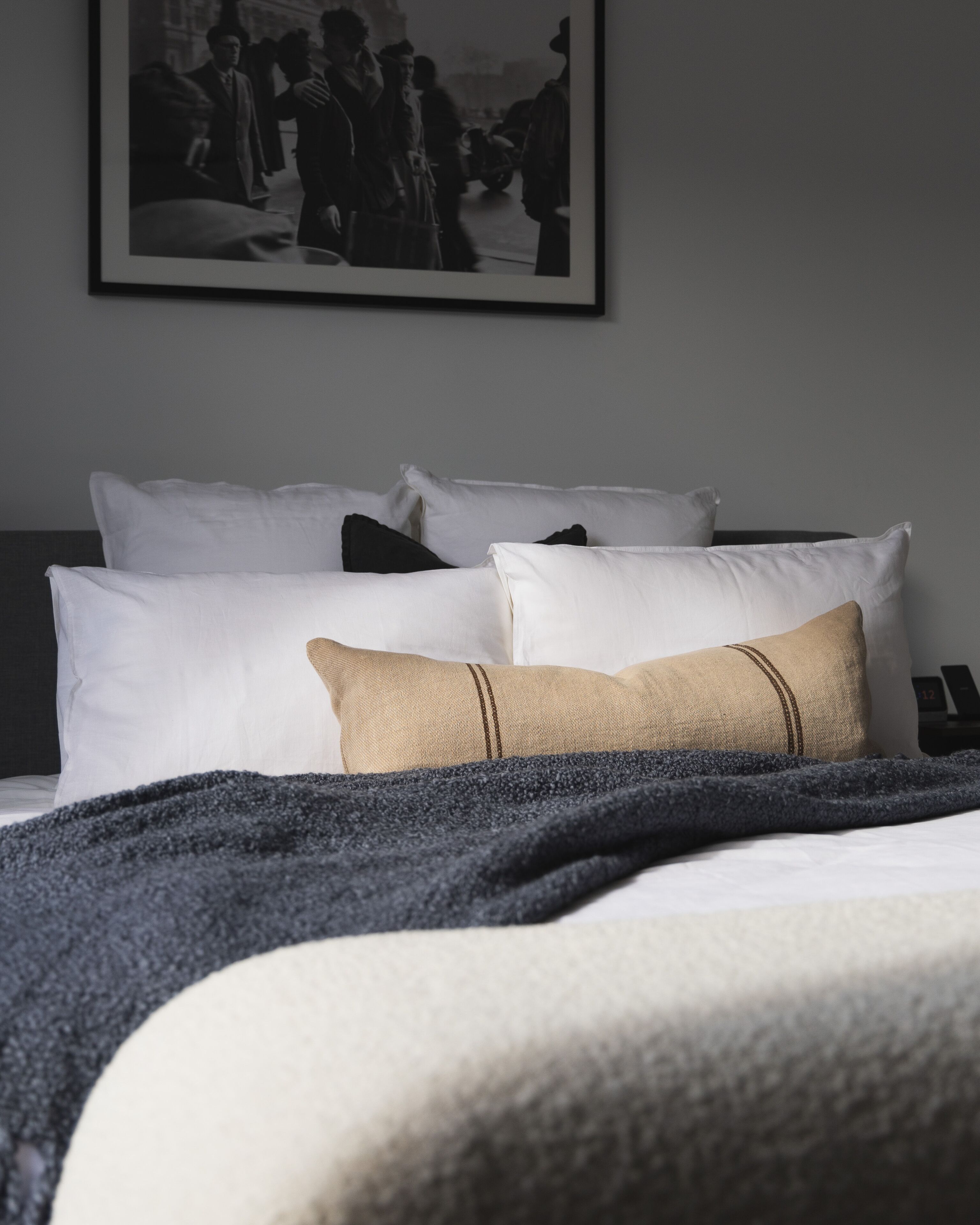 Un letto rifatto con cuscini bianchi e beige, una coperta scura e una fotografia in bianco e nero incorniciata al muro.