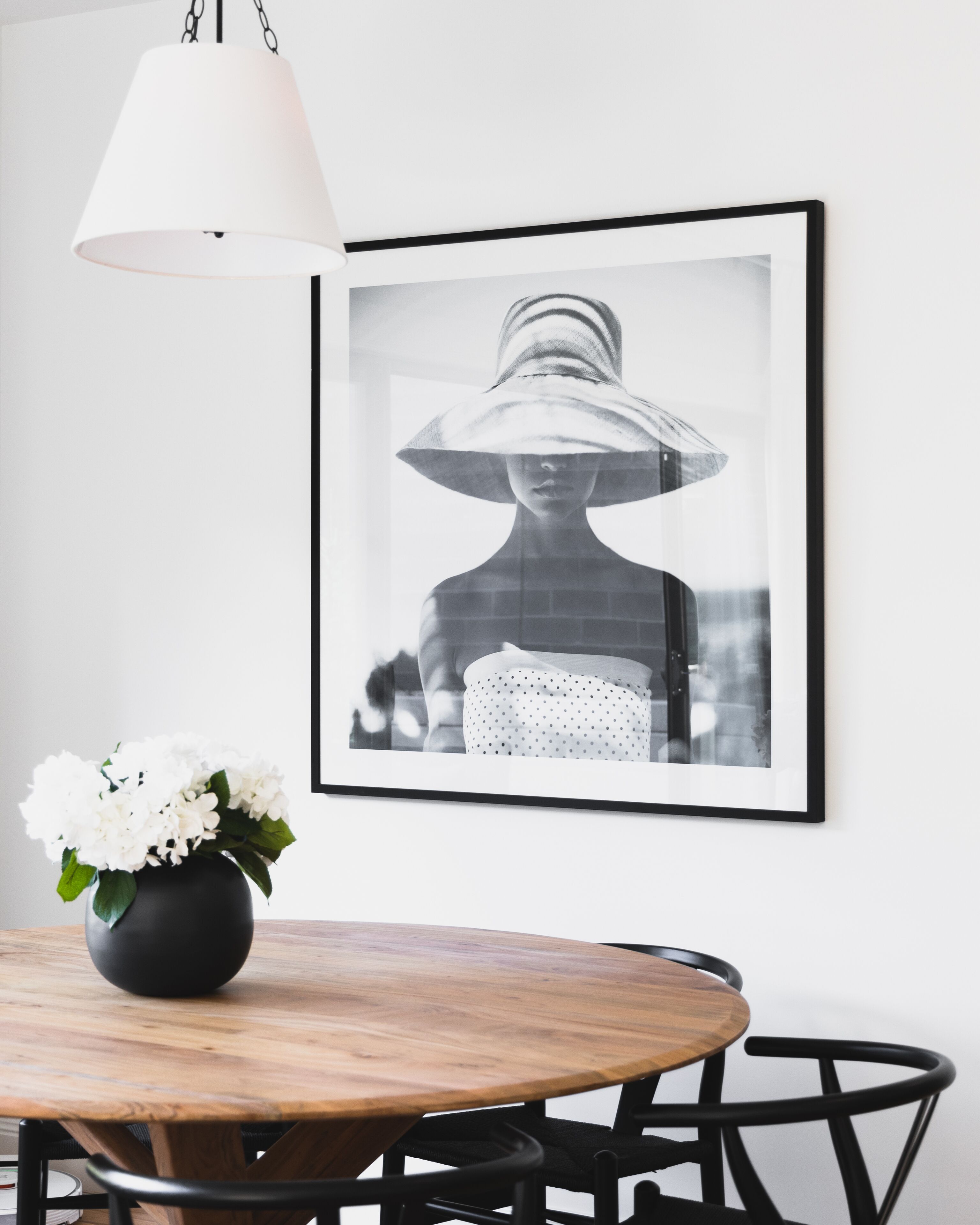 A dining area with a round wooden table, black chairs, a black vase with white flowers, and a framed black and white photograph on the wall.