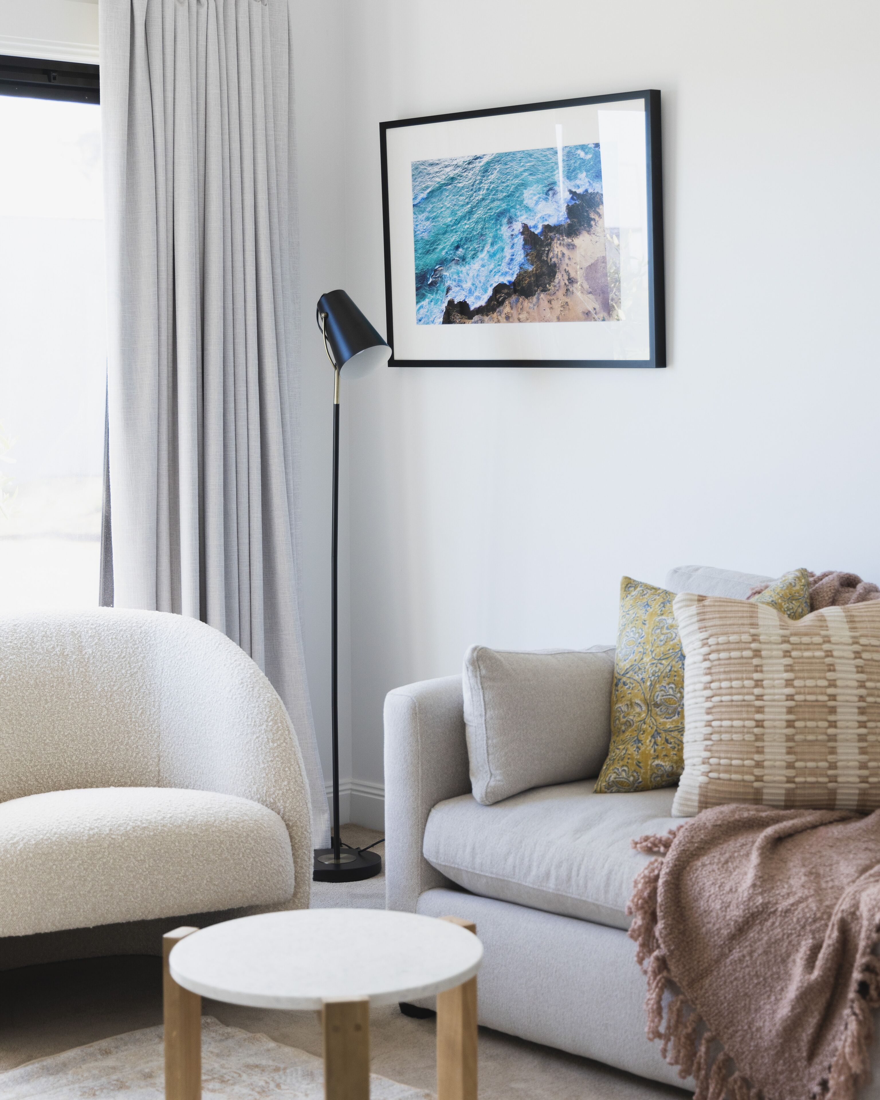 Un salon moderne avec un canapé, une table basse ronde et un lampadaire. Un tableau encadré représentant une scène de plage est accroché au mur.
