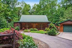 A rustic log cabin with a green roof, surrounded by lush greenery and a paved driveway.