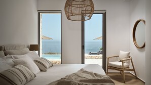 Een slaapkamer met een groot bed, een stoel, een hanglamp en uitzicht op het strand door een schuifdeur.
