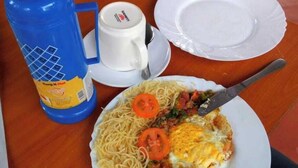 A meal with spaghetti, a fried egg, and a tomato slice on a plate, a fork, a knife, a white cup, and a blue water bottle on a wooden table. - Savannah Road (Nairobi)