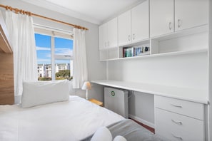 A bedroom with a bed, white curtains, a window with a view, a desk with books, and a radiator.