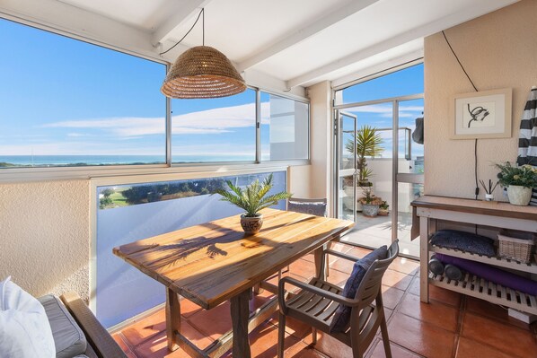 A balcony with a wooden table, chairs, a hanging light fixture, and a view of the ocean.