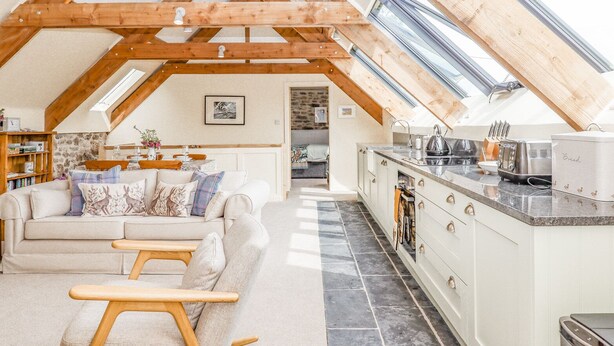 A cozy loft-style living space with a high ceiling, exposed wooden beams, a kitchen area, and a dining area. - The Coach House at Thorn Farm (Exeter)