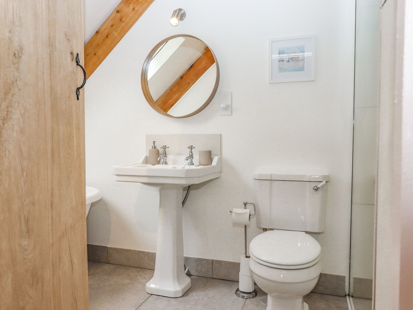 Un baño con lavamanos pedestal, inodoro, espejo redondo y un cuadro en la pared.