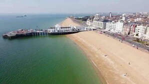 Een kustgebied met een zandstrand, een pier die in het water uitsteekt en gebouwen op de achtergrond.