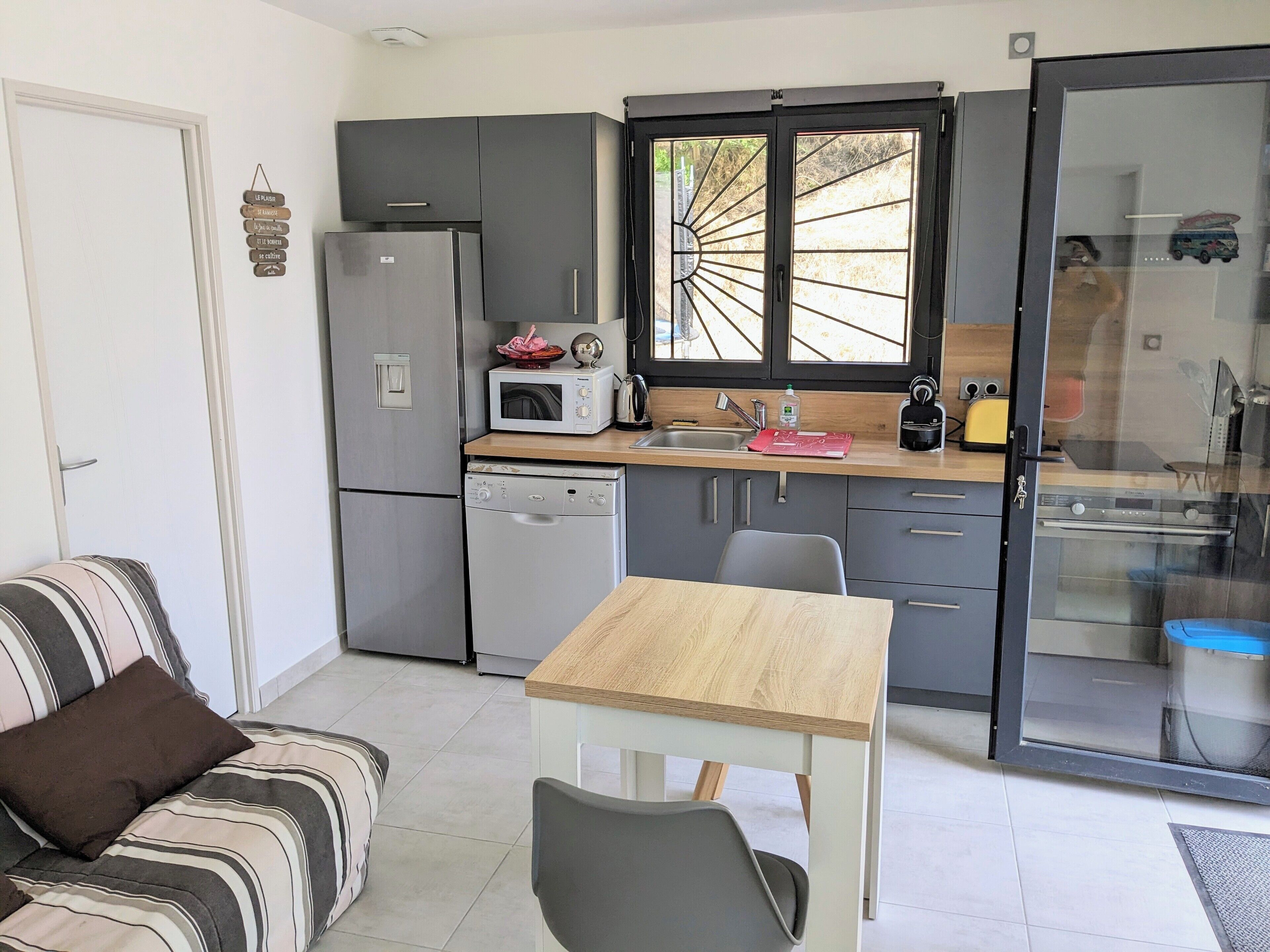 Una cucina compatta con frigorifero, microonde e lavastoviglie. C'è un tavolo da pranzo con sedie, un divano a righe e una porta vetrata scorrevole che dà su un balcone.