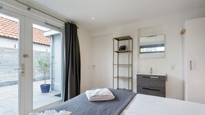 A hotel room with a bed, a small desk, a bookshelf, a mirror, and a window with a view of the outside.