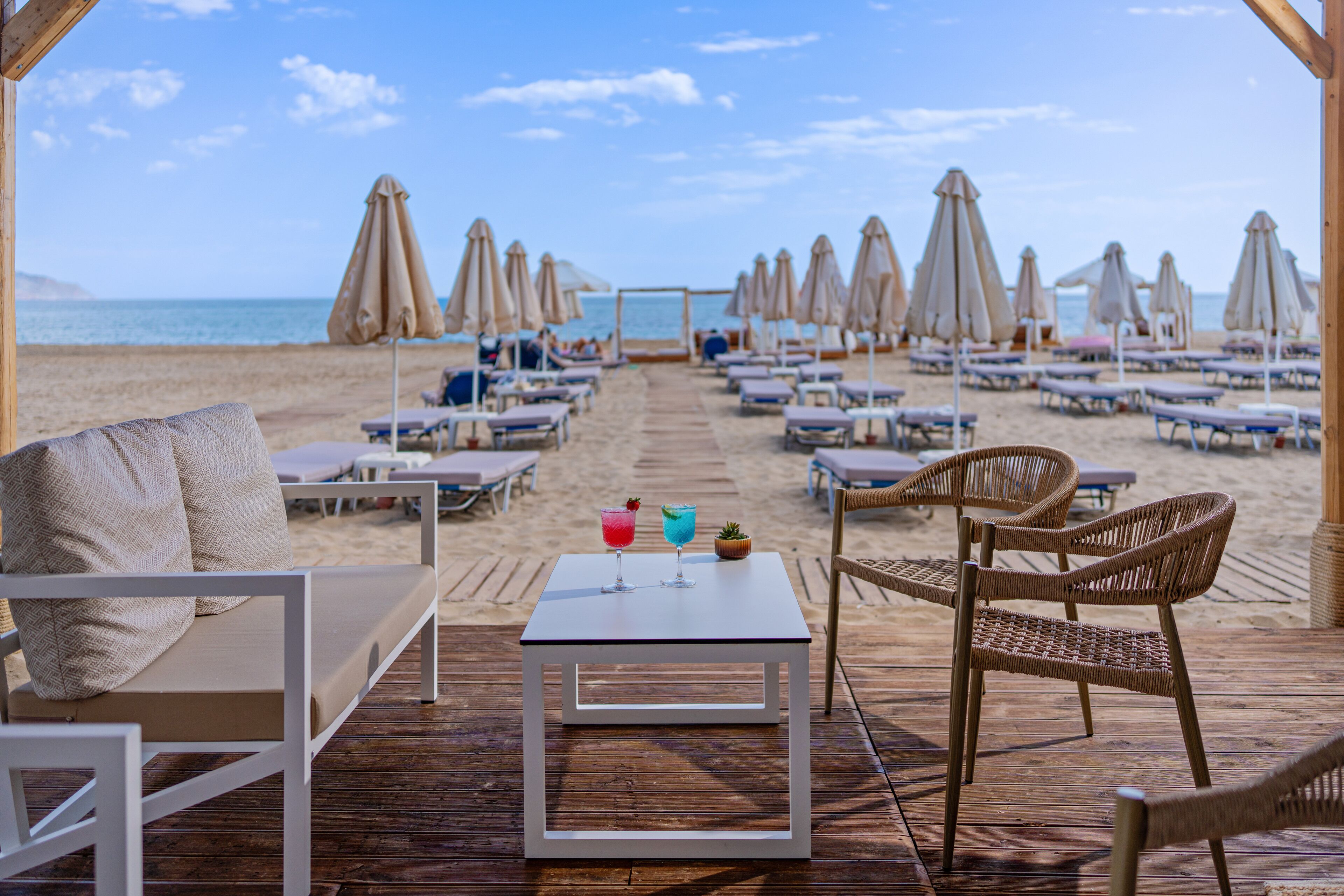 A beachside lounge with outdoor seating, a table with drinks, and umbrellas providing shade.