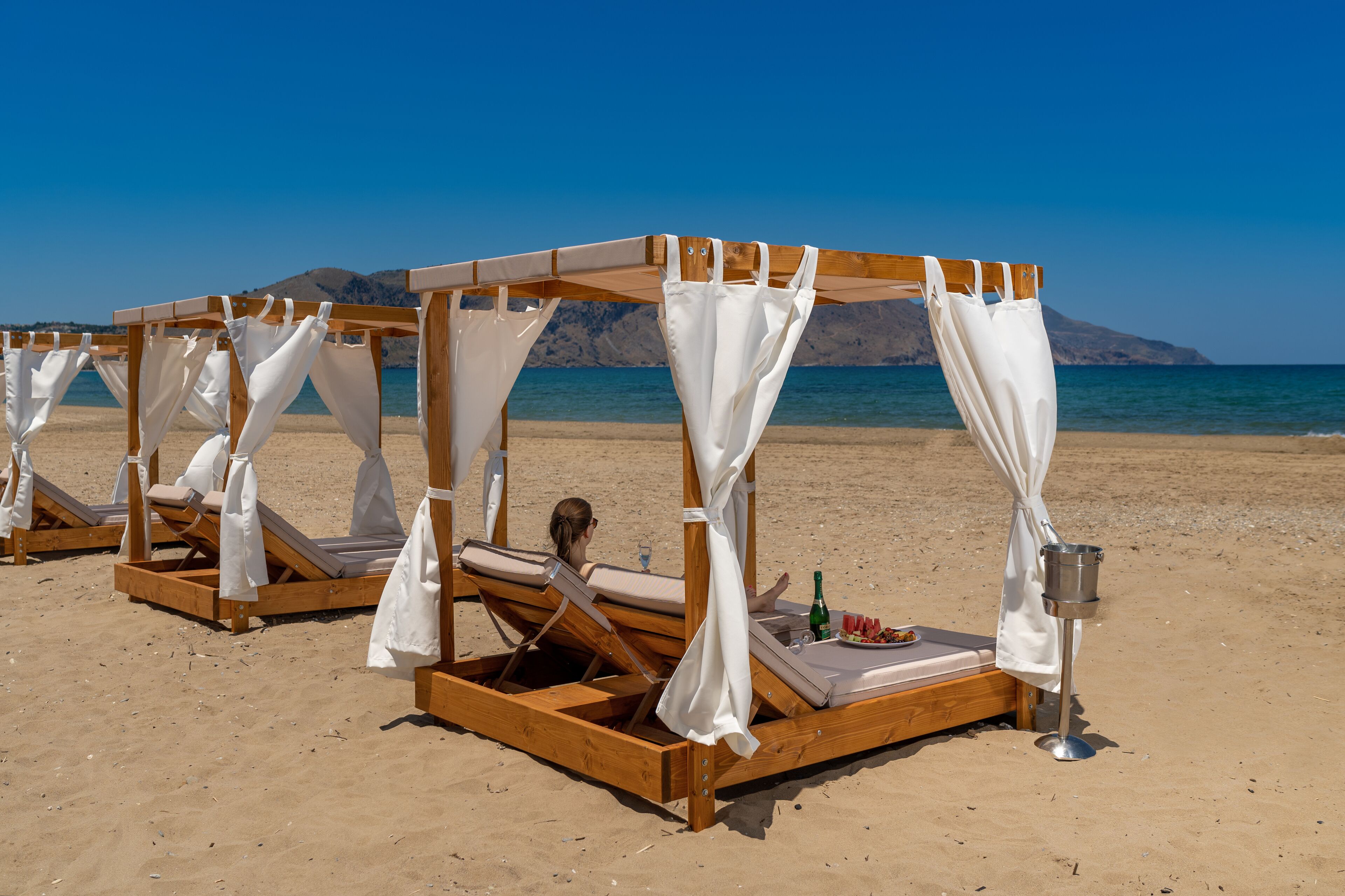 A beach with wooden canopy beds, a bottle of champagne, and a plate of food.