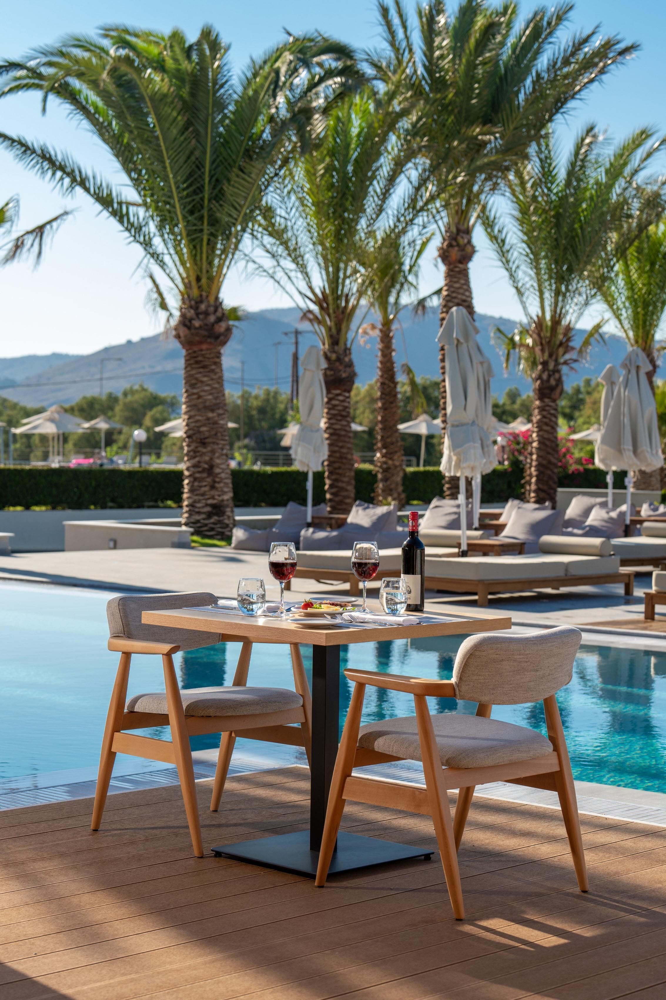 A poolside dining area with a table set for two, surrounded by palm trees and lounge chairs.