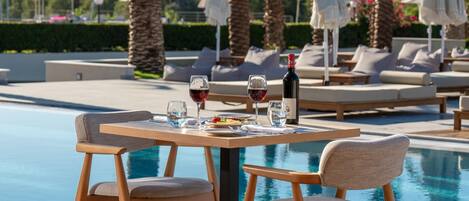 A poolside dining area with a table set for two, surrounded by palm trees and lounge chairs.