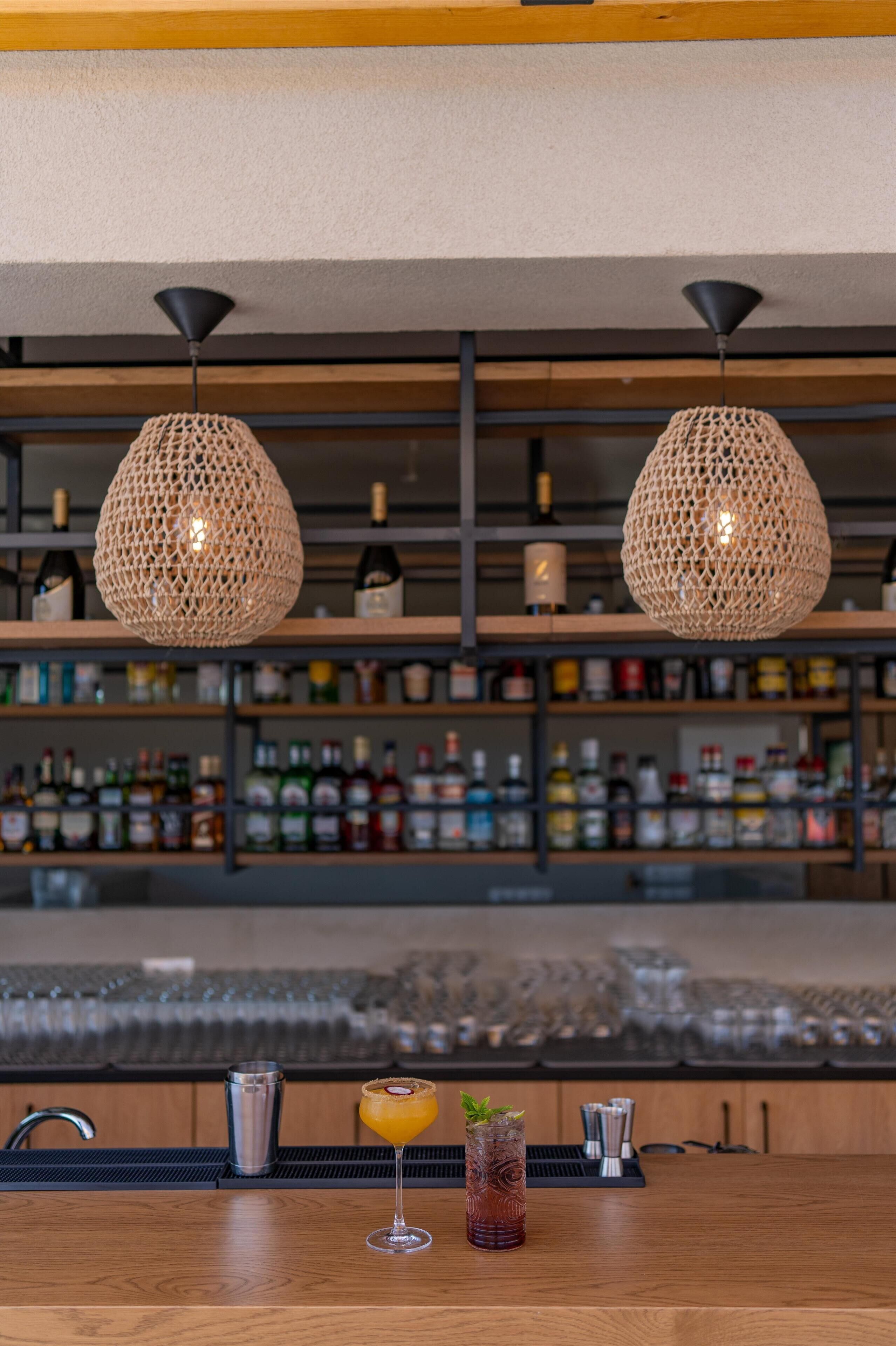 A bar area with two woven pendant lights, a wooden bar counter, and a variety of bottles in the background.