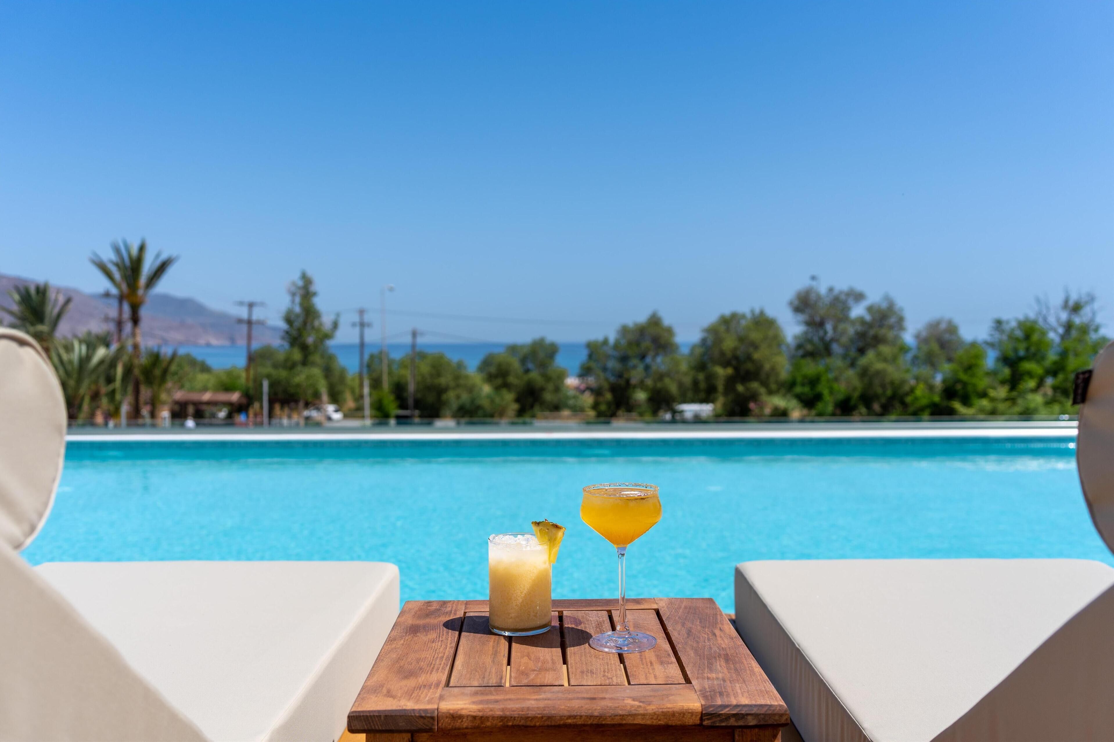 Ein Holztisch mit zwei Getränken und einem Glas, mit Blick auf ein klares, blaues Schwimmbecken und einen Blick auf das Meer und das Grün im Hintergrund.